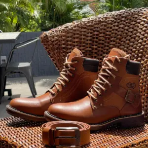 Par de botas de cuero marrón para hombre, estilo trabajo con cordones claros, hebilla lateral y cinturón a juego, sobre silla de mimbre al aire libre.